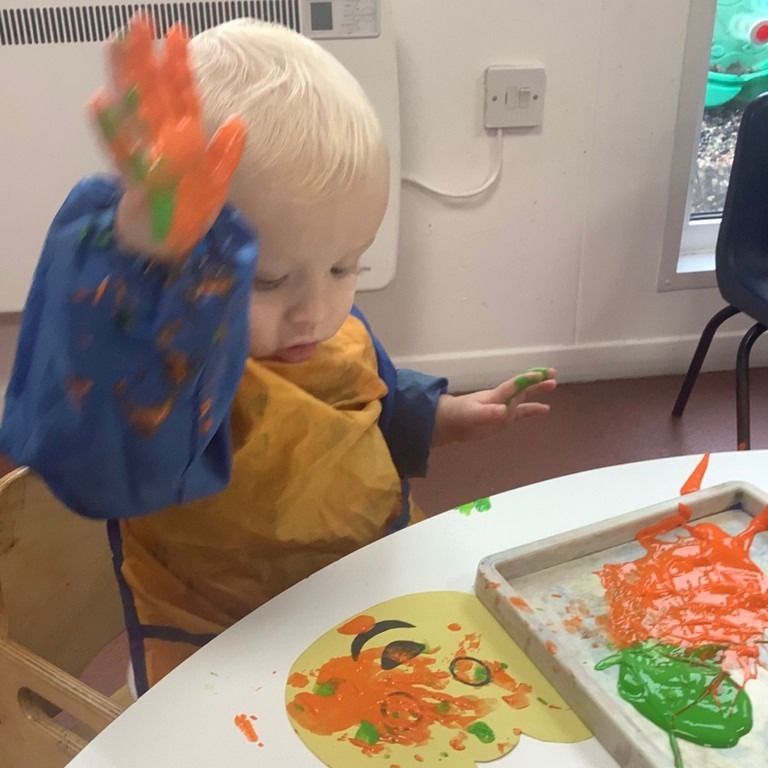 A toddler in an orange paint apron, hands covered in orange and green paint, finger painting at a table