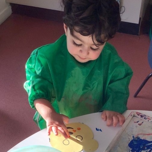 A toddler in a green paint apron studying their paint-covered hand at the painting table