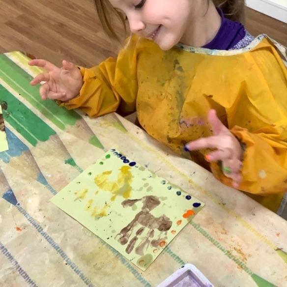 A child in a yellow paint apron smiling while painting a horse with their fingertips