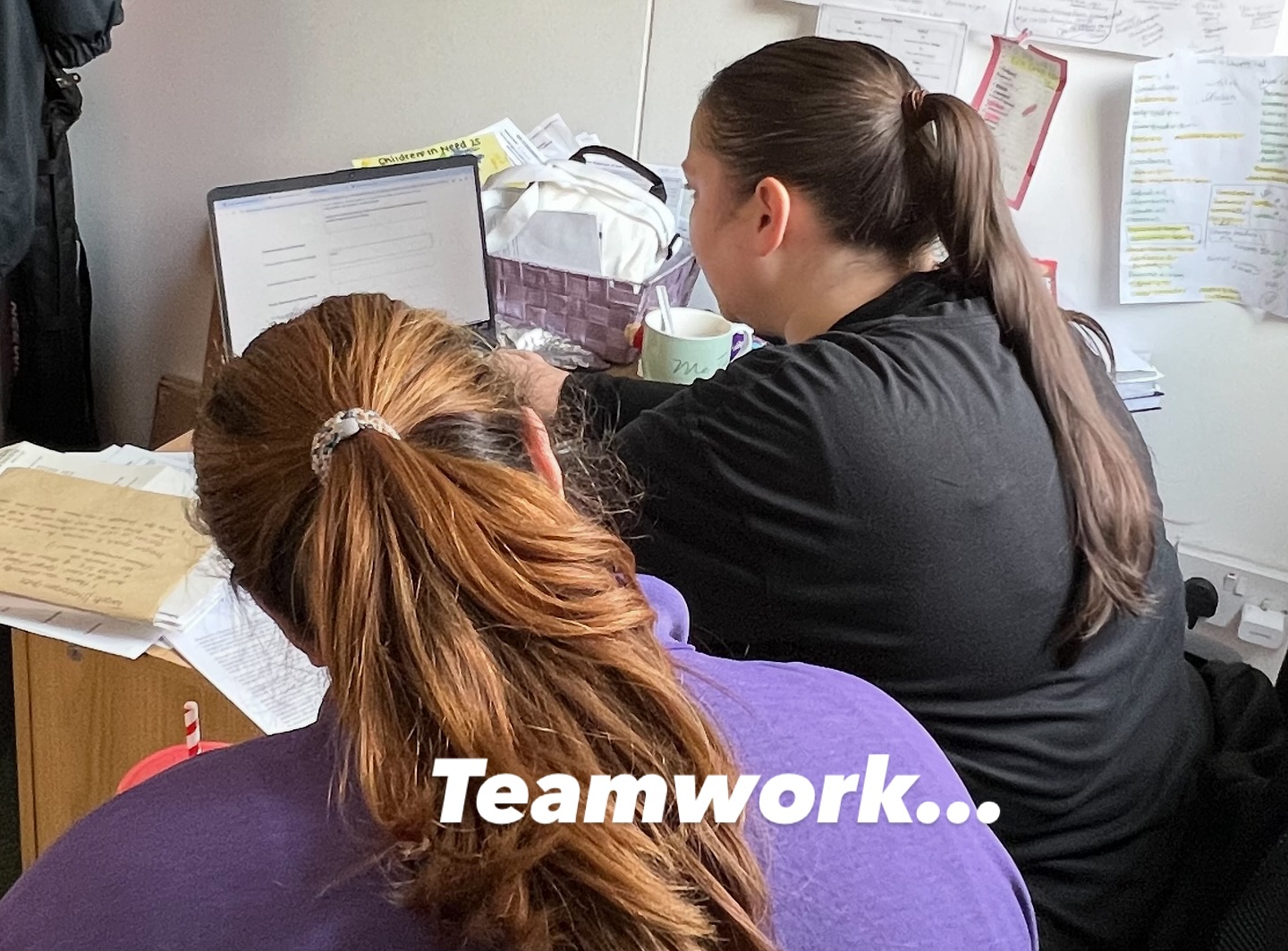 Two staff members working together at a desk planning the day — paperwork, laptops, mugs of tea